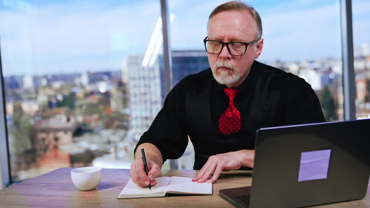 Busy entrepreneur looks through his paper notebook. Pensive businessman looks at laptop and drinks from cup.