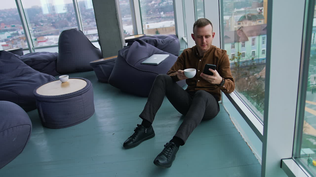 Young businessman having break. Handsome guy in shirt and trousers sitting on a floor with a cup of coffee and a phone near the window in modern office in the city.