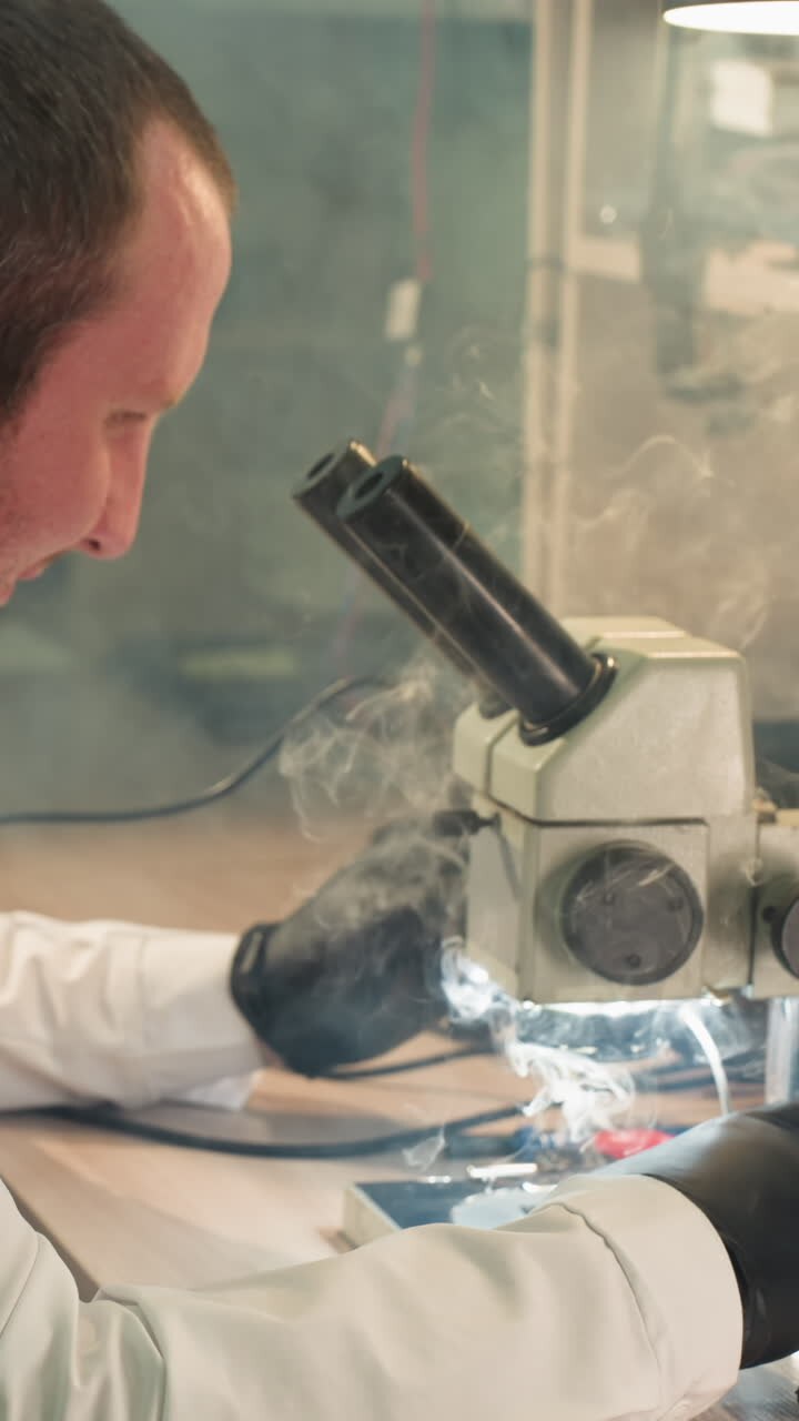 A close-up view of a technician in a white lab coat working on an electronic circuit board under a microscope, with smoke from the soldering iron fill the place