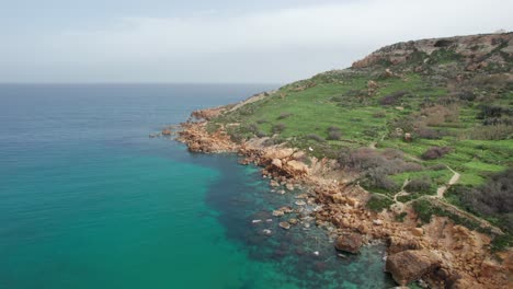 Flying-Over-A-Orange-Rocky-Shoreline-With-Turquoise-Water-At-The-Base-Of-A-Green-Hill