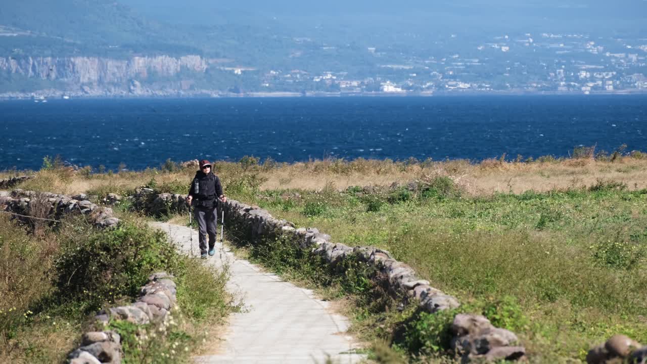 Seorang pejalan kaki menjelajahi Jalur Olle yang menawan di Pulau Jeju. Dikelilingi oleh dinding batu tradisional dan lahan pertanian subur, jalur ini menawarkan pemandangan laut yang menakjubkan di bawah langit cerah.