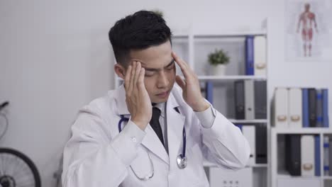 slow motion with shoulder shot asian physical suffering migraine is rubbing his temple in circular motion while taking a break at a white medical workplace.