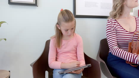 Caucasian-girl-patient-using-tablet-sitting-with-mother-in-hospital-reception,-slow-motion