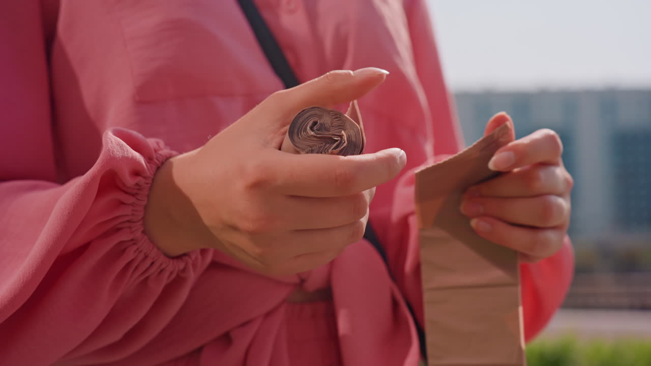Casual Woman Retrieving Chocolate During Walk, Woman Opening Small Chocolate Coin Outside Near City Skyline, Casual Woman Unwraps Chocolate Coin From Foil While Walking Outside With City View