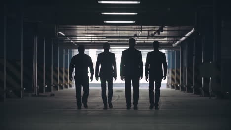 slow motion locked down rear view of silhouettes of four men in black suits confidently walking away from camera towards exit of parking lot