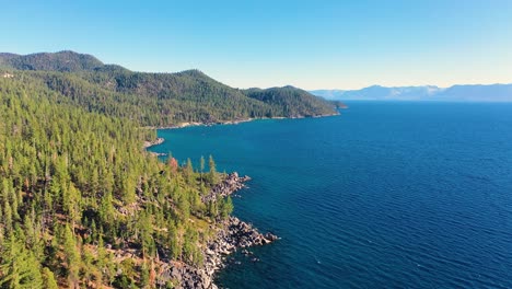 toma aérea de agua con agua azul cristalina y espeso bosque de pinos con cordillera y costa rocosa en el lago tahoe, california