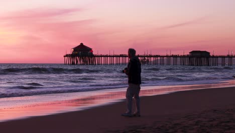 a man takes a picture of the beach during a gorgeous yellow, orange, pink and blue sunset with the huntington beach pier in the background at surf city usa california