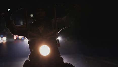 biker riding his chopper motorcycle with high handlebars on a highway at night in the rain. viewed from his front in close-up, silhouetted by bright headlights of a car behind him in background