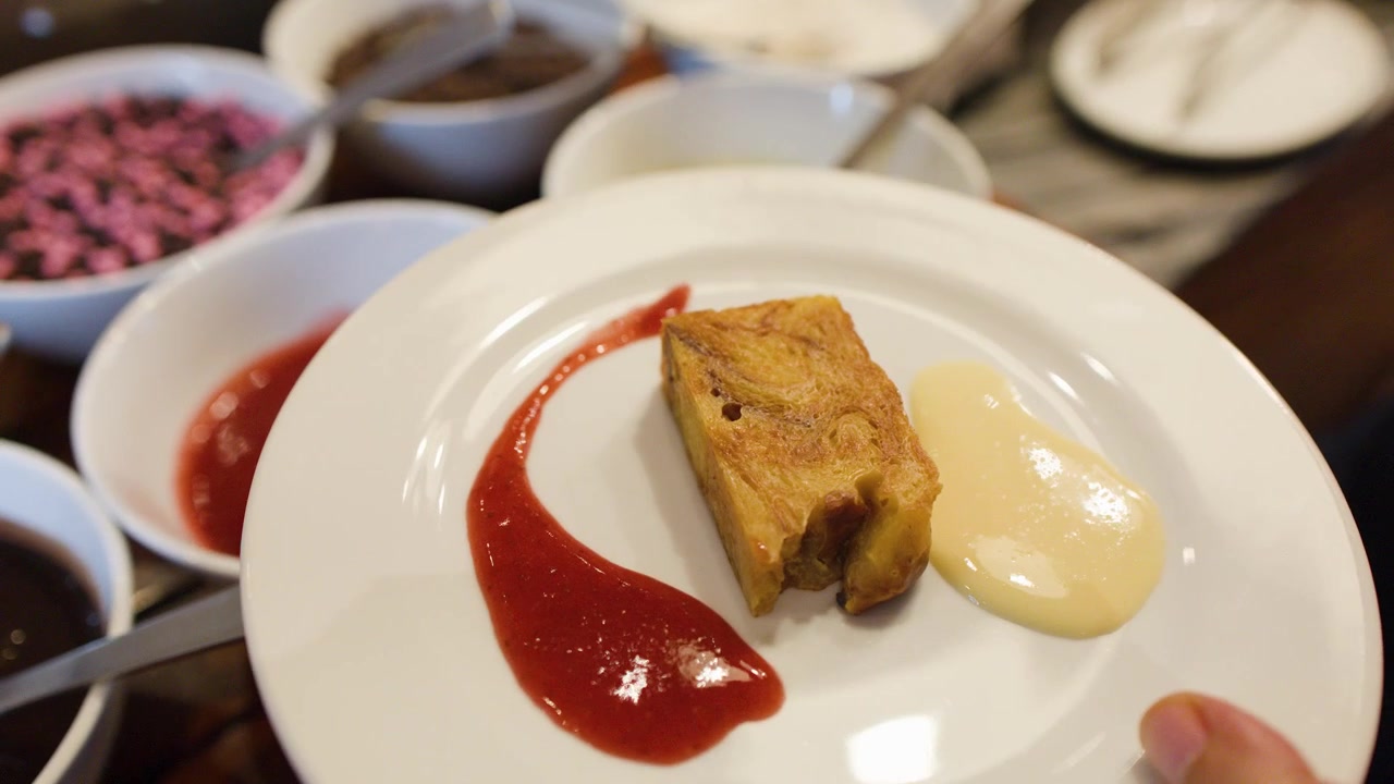 Een metalen lepel giet rode aardbeiensaus op een wit bord met broodpudding dessert
