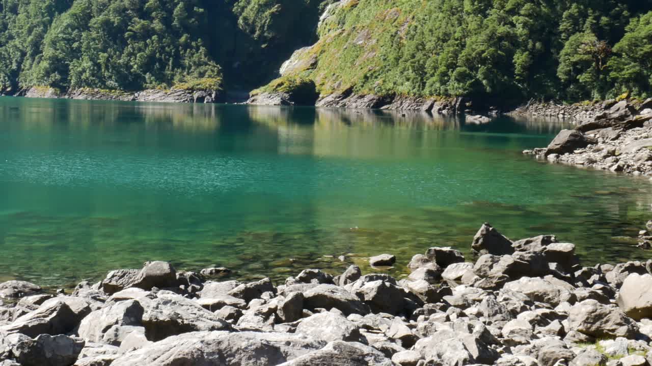 뉴질랜드의 바위가 많은 해안선과 함께 햇빛에 의해 열대 호수 마리안 조명의 패닝 샷