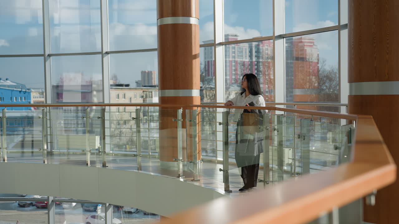 Young professional marketer dressed in business casual attire leans gently on wooden mall railing, gazing down with calm focus, surrounded by large glass windows and urban skyline backdrop