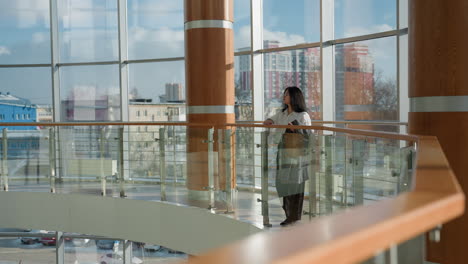 young professional marketer dressed in business casual attire leans gently on wooden mall railing, gazing down with calm focus, surrounded by large glass windows and urban skyline backdrop