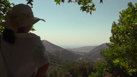 Back-of-girl-holding-on-to-summer-hat-while-looking-out-onto-distant-mountain-landscape-framed-by-trees-SLOW-MOTION
