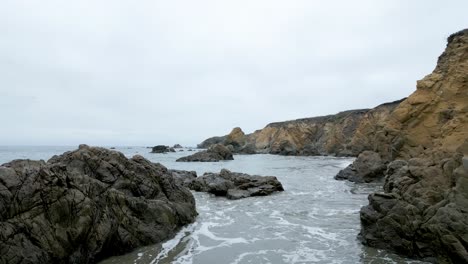 Volando-Bajo-Sobre-El-Océano-En-La-Playa-Del-Condado-De-San-Mateo-Sobre-Rocas-Y-Olas,-California