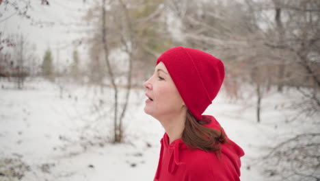 close up of sports woman wearing red beanie and hoodie jogging outdoors during winter surrounded by snow-covered trees, serene park pathway, evergreen pines, and foggy atmosphere