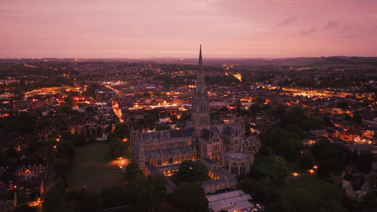 Veduta aerea della Cattedrale di Salisbury al tramonto con un cielo crepuscolare rosa, strade cittadine illuminate e la storica cattedrale che si erge sopra Salisbury