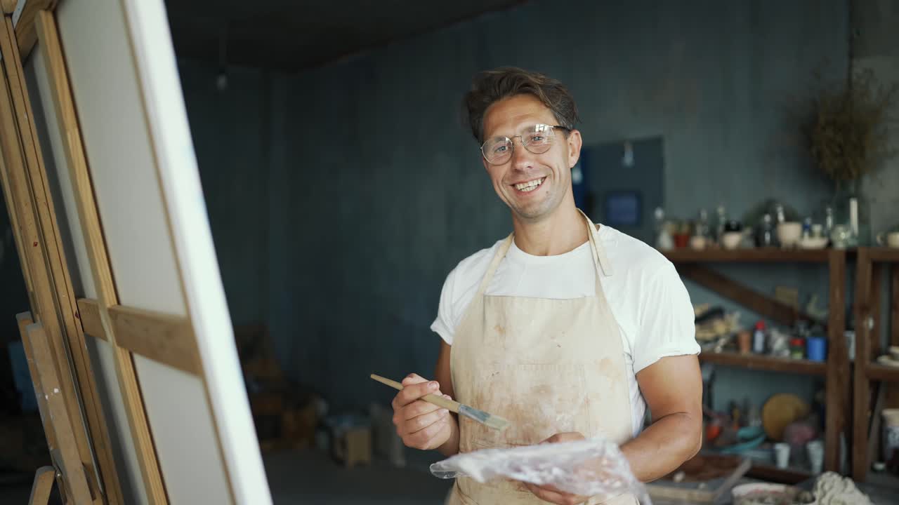 Portrait of a young happy artist in glasses with a brush near the canvas. The man smiles and poses. Creative everyday life