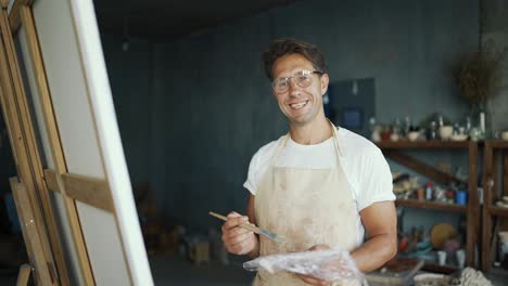 Portrait-of-a-young-happy-artist-in-glasses-with-a-brush-near-the-canvas.-The-man-smiles-and-poses.-Creative-everyday-life