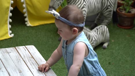 adorable one-year-old baby girl posing for pictures in outdoor camping site installation near home on the grass leaning on the small wooden table wearing jean dress and bow