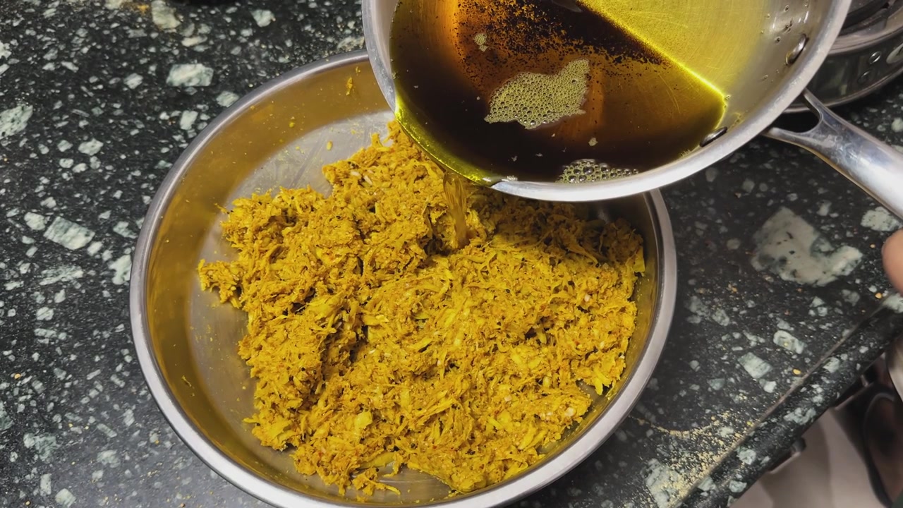 Close view of traditional crushed mango pickle preparation with hot mustard oil being poured over spiced shredded mango mixture inside a steel bowl during homemade Indian achar making process