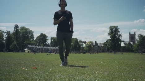 joven blanco caminando sobre la hierba mientras revisa su teléfono móvil en un parque de la ciudad de cambridge, inglaterra