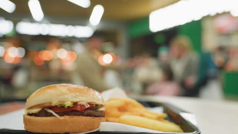 patatas fritas doradas y una hamburguesa en una bandeja negra en una mesa con personas borrosas en el fondo, destacando la deliciosa comida rápida en un ambiente de comedor animado