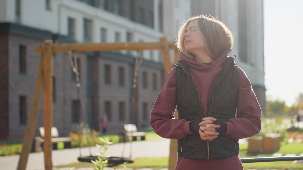Fit lifestyle advocate closes eyes and gently rotates neck in warm up exercise beside urban workout bars under bright sunshine, wearing hoodie and vest, peaceful outdoor fitness moment