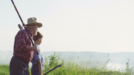 Side-view-of-a-grandfather-and-grandson-holding-their-fishings-rods-and-talking-on-the-shore-of-the-river