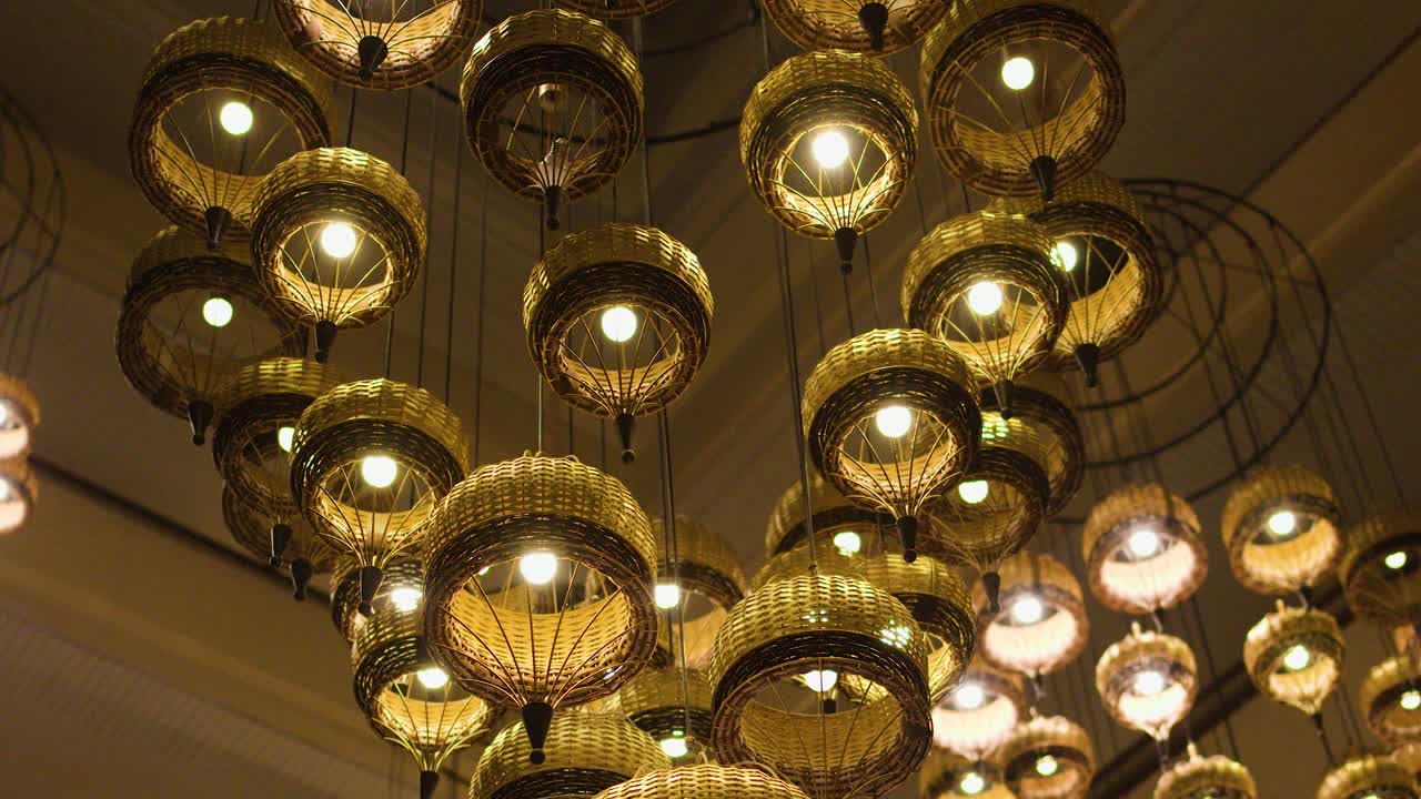 Warm wicker chandeliers glow in elegant Thai hotel, camera pans upward revealing intricate lighting design