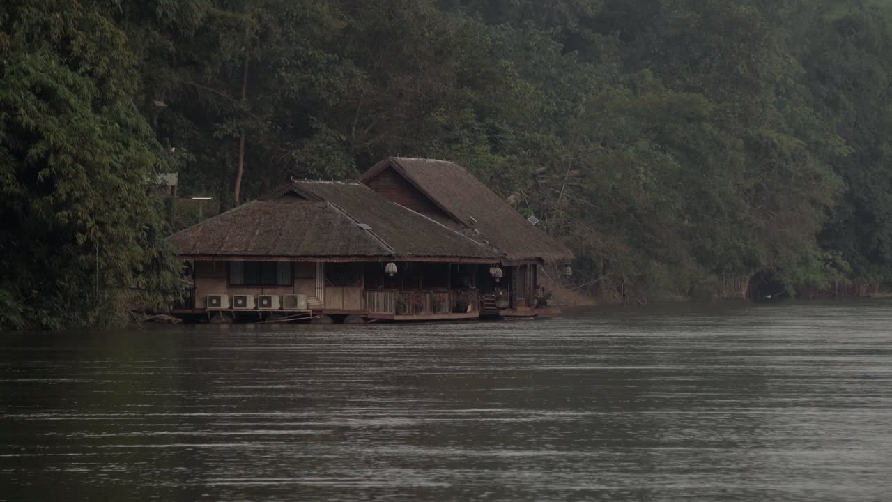 una toma distante de una casa flotante tailandesa en la orilla del rápido río khwae noi en kanchanaburi, tailandia