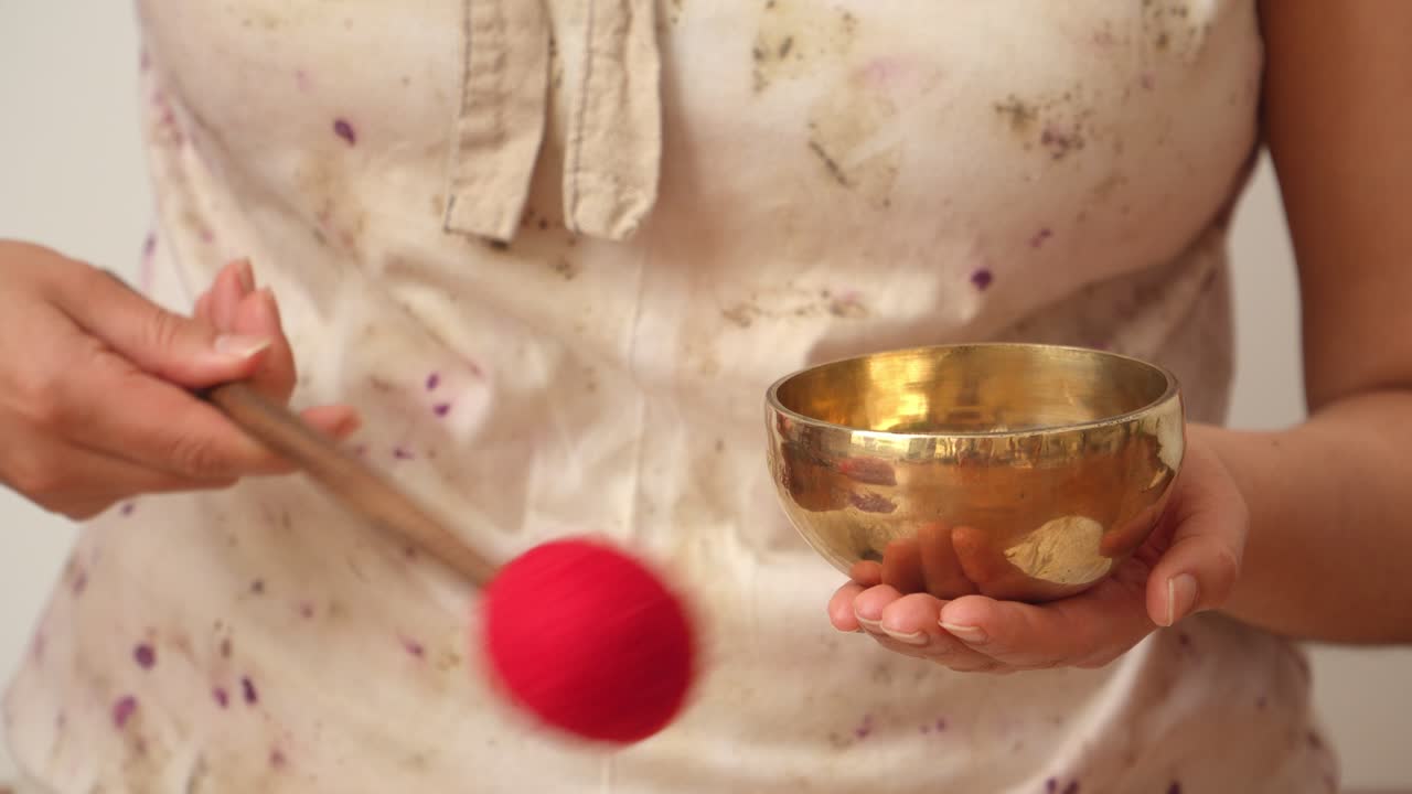 Medium close-up of a woman holding and ringing a small Tibetan singing bowl in her hand inside a calm minimalist indoor studio