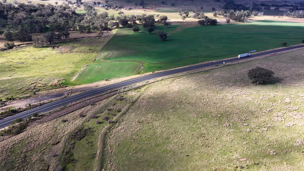 A white semi-truck travels along a winding rural road through grassy fields and farmland, captured by a moving aerial drone under natural daylight