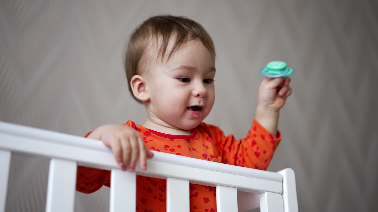 Beautiful Caucasian baby boy stays up in white cot. Cute kid playing with pacifier throwing it on the floor and watching it fall. Low angle view.