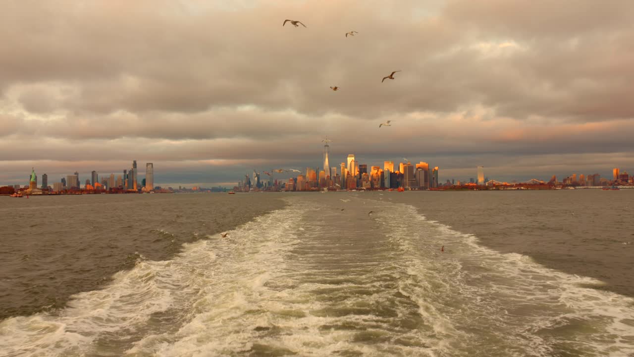 A ferry moves on the Hudson River with a view of the Manhattan skyline in the background. Gray clouds and city lights create a dynamic atmosphere. This shot captures urban life in New York City