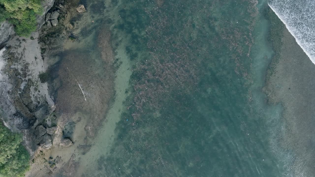 Top down quickly descending drone shot of polluted water filled with trash floating over dead coral reef in the turqouise tropical water of Bali Indonesia