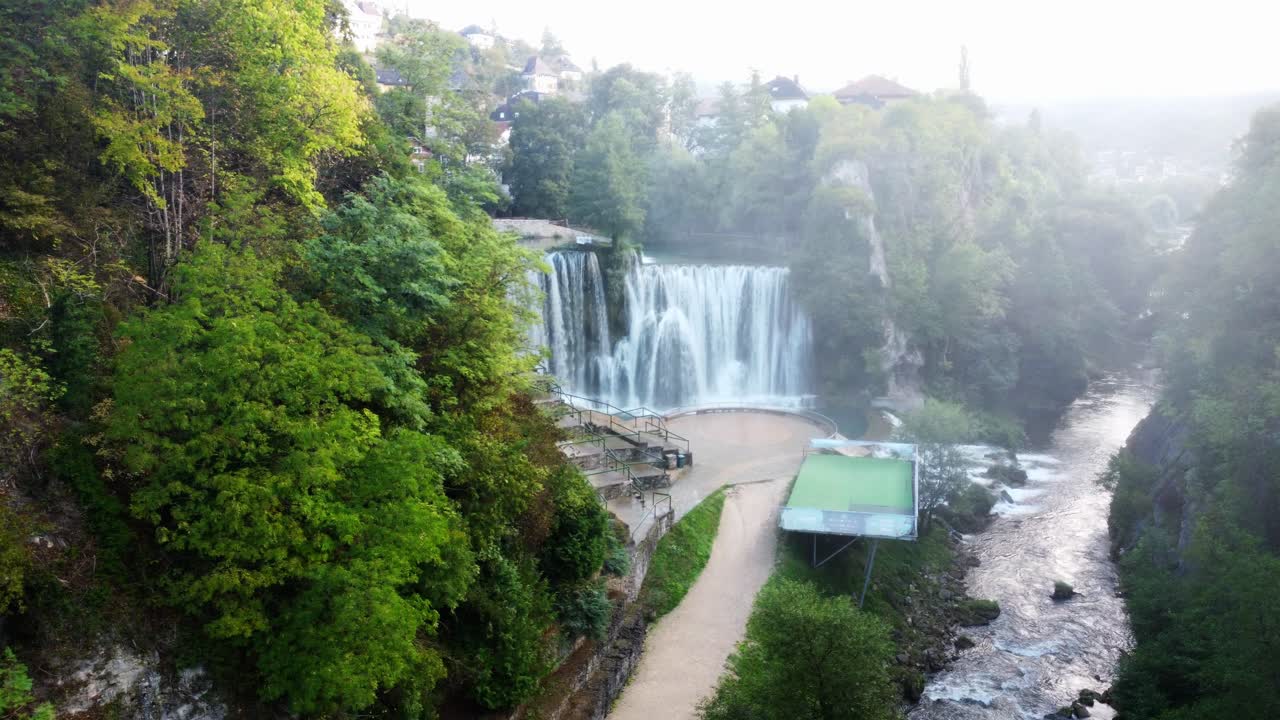 Filmato aereo della cascata di Jajce all'alba in Bosnia ed Erzegovina. Veduta aerea della cascata della città circondata dal verde, nebbia mattutina e luce calda, ideale per progetti di viaggio e turismo.