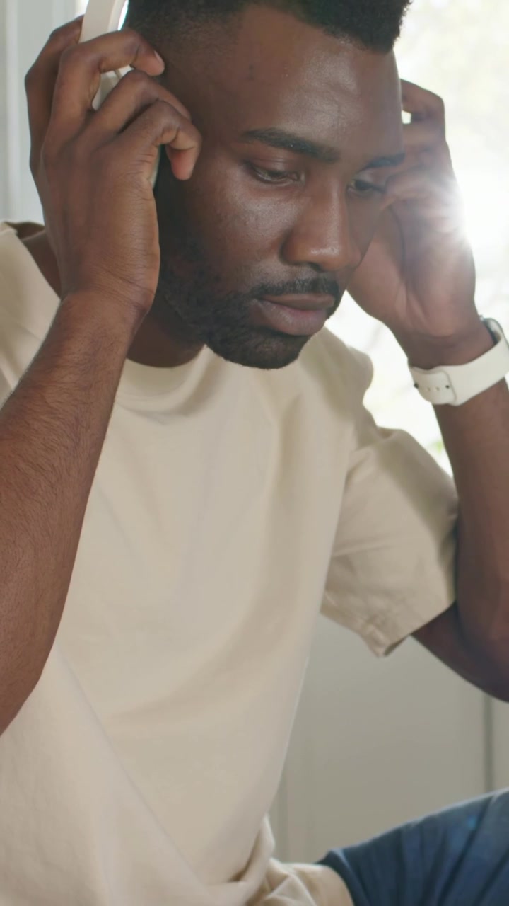 Vertical video: Adjusting white headphones man wearing beige tee checking phone at kitchen table