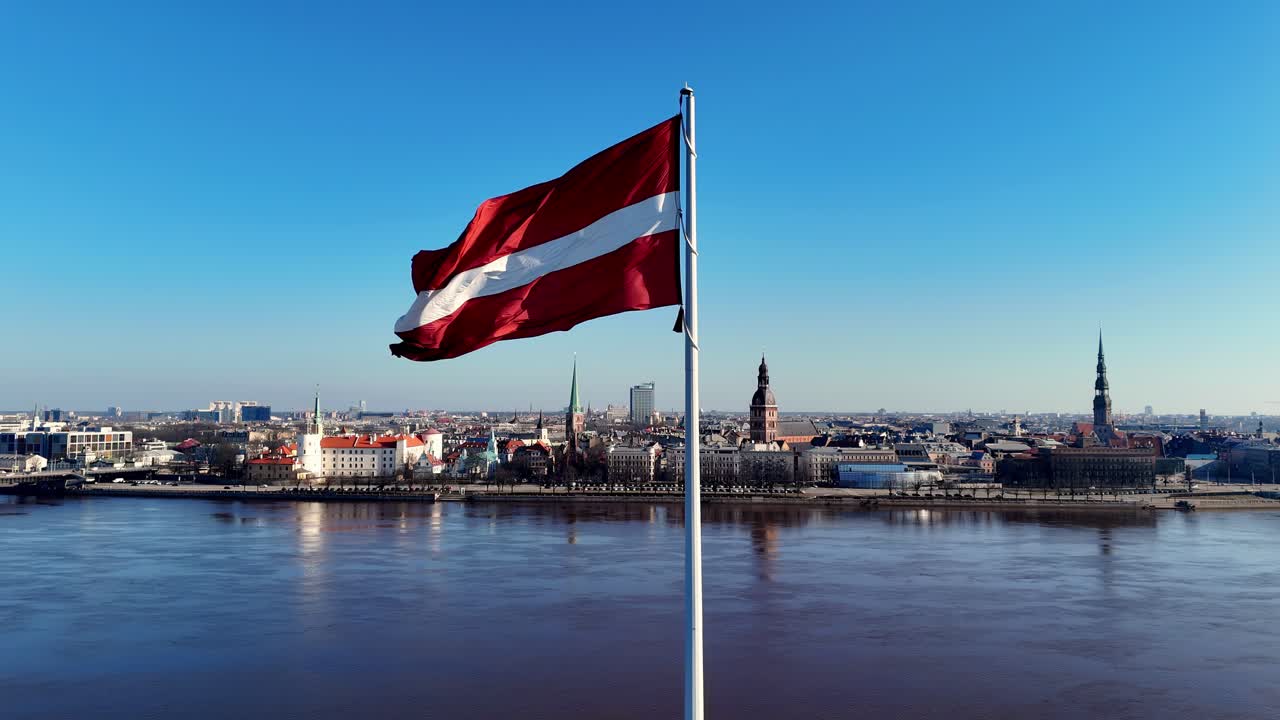 A waving Latvian flag on a flagpole in front of a Riga cityscape with modern high-rise buildings made of glass and metal