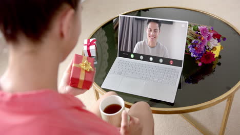 mujer caucásica sosteniendo un regalo rojo usando una computadora portátil con un hombre caucásico en la pantalla