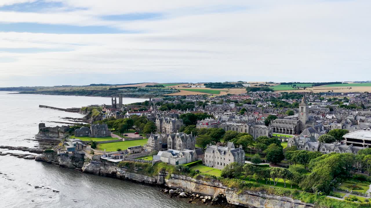 Drone glides over St Andrews coastline, castle ruins, cathedral, university buildings, and dramatic cliffs