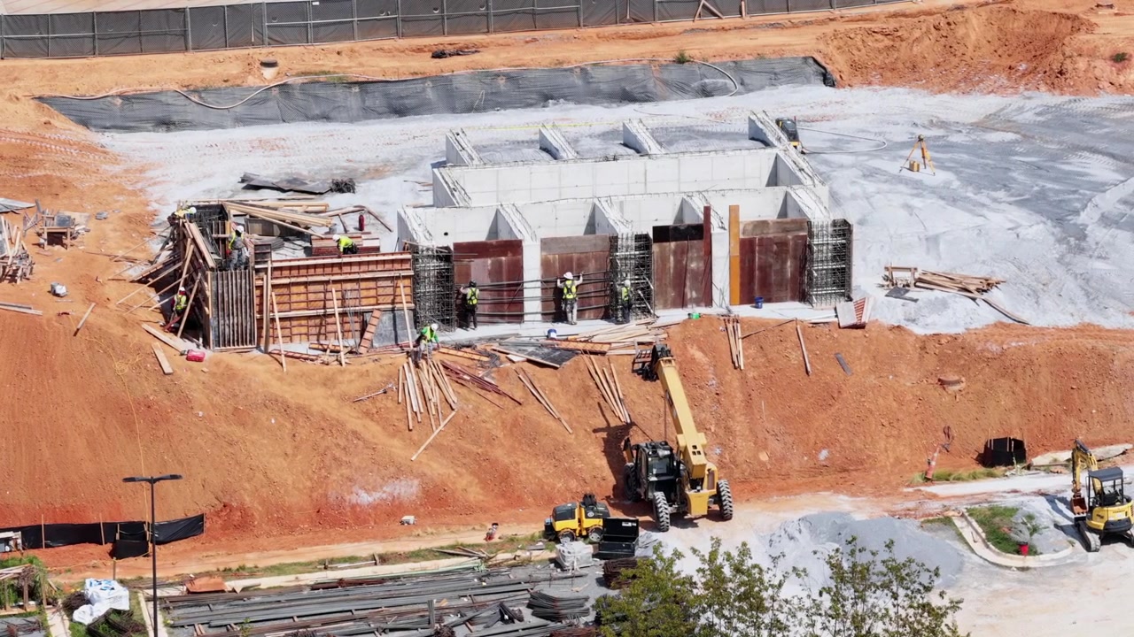 Workers in safety vests build concrete retaining wall near Old Graves Mill Road, telehandler lifts materials across red clay excavation, wooden forms line foundation site. Aerial drone view.