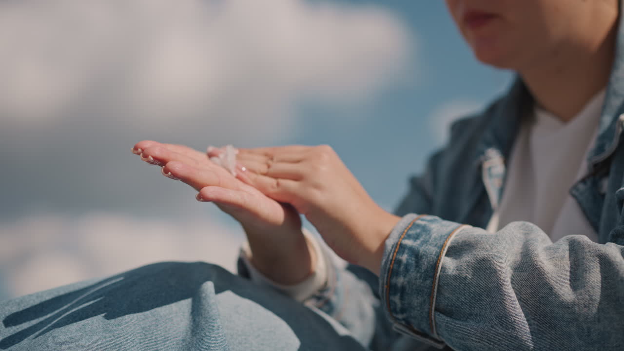 Sunscreen on hands under blue sky, young woman in denim jacket rubs sunscreen into palms during breezy picnic with clouds overhead, closeup texture of lotion and protective skincare routine