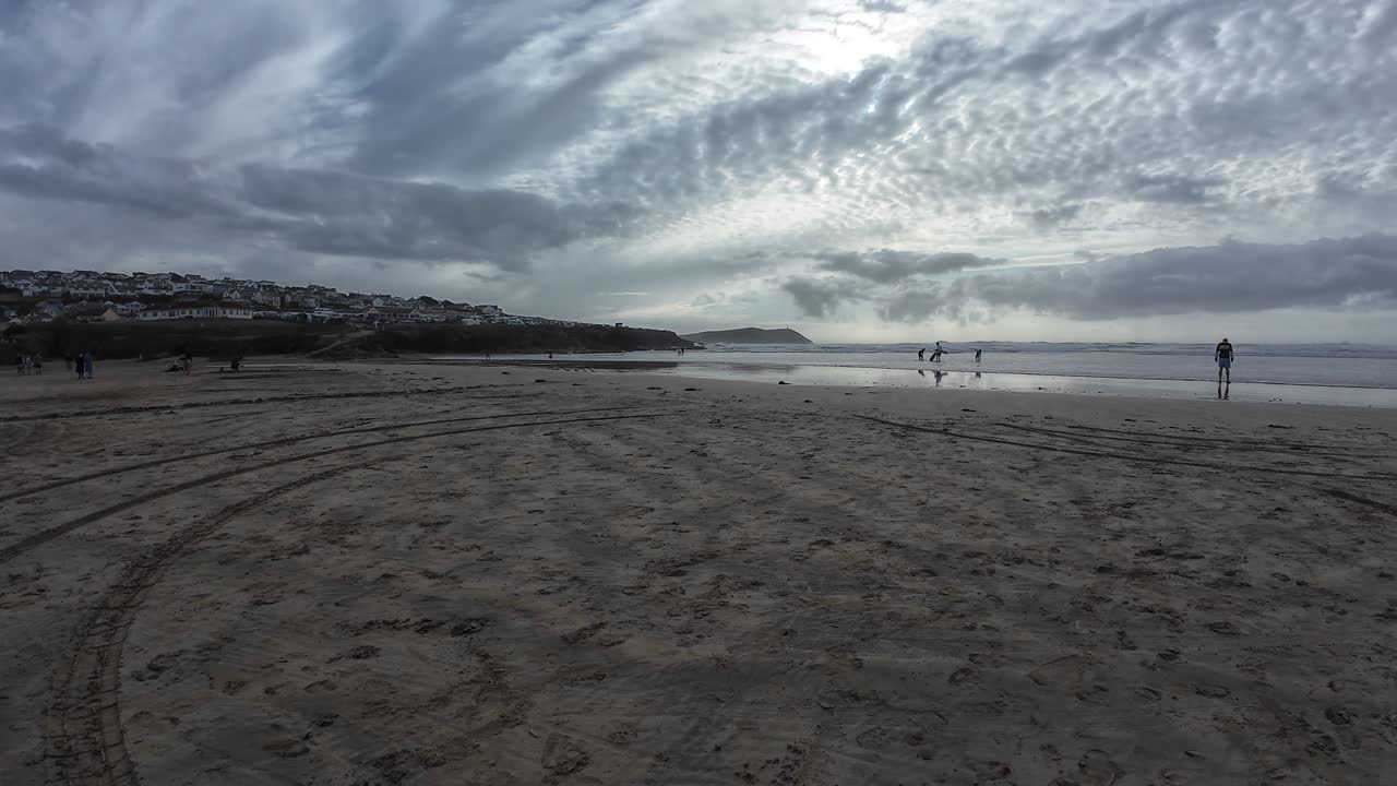 A tranquil view of Polzeath Beach in Cornwall, UK, with artistic sand circles in the foreground, people walking by the shore, and a dramatic cloudy sky over the Atlantic coastline