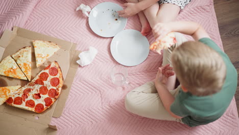 overhead view of two little kids sitting on soft pink blanket on wooden floor eating slices of pepperoni and cheese pizza from open box with empty plates and napkins scattered around