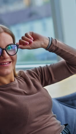 happy smiling lady touching her glasses. relaxed woman sitting in bean bag chair resting from work. blurred backdrop. vertical video