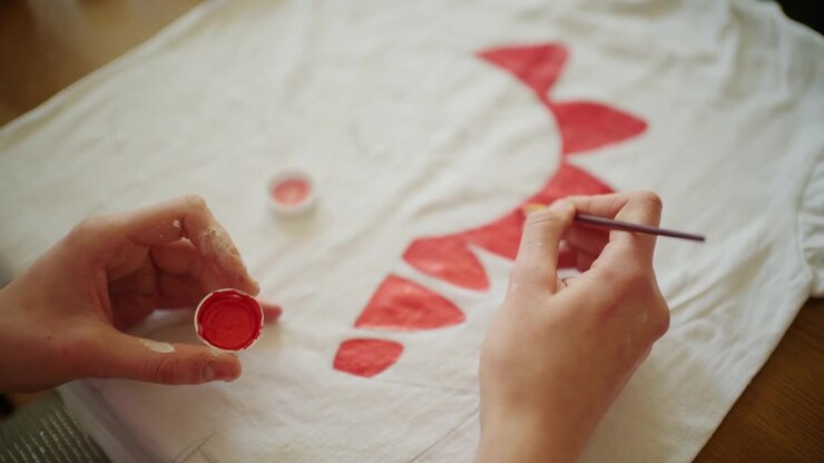 Primo piano di un artista che dipinge forme decorative rosse su una maglietta bianca usando pittura per tessuti