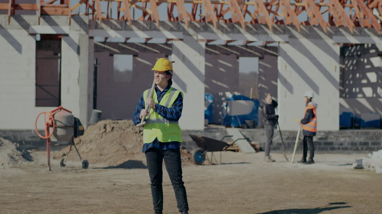 Young male worker wearing hardhat and reflective clothing carrying shovel on shoulder while standing at construction site on sunny day