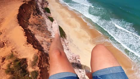 Piernas-De-Un-Hombre-Sentado-En-Una-Silla-De-Un-Parapente-Con-Acantilados-De-Arena-Y-Hermosas-Aguas-Azules-Del-Océano-Debajo-En-La-Ciudad-Tropical-De-Pipa-En-Rio-Grande-Do-Norte,-Brasil-En-Un-Cálido-Día-Soleado-De-Verano