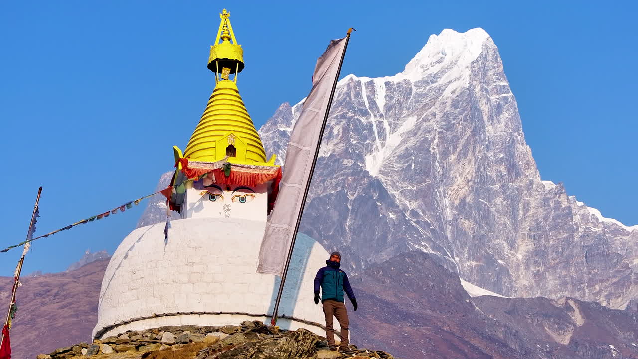 A male tourist waves to drone shot at a Buddhist stupa in Dingboche, Khumbu region, Nepal. Mt. Cholatse rises in backdrop with Buddhist flags, blue sky, and scenic views on the Everest trek serenity