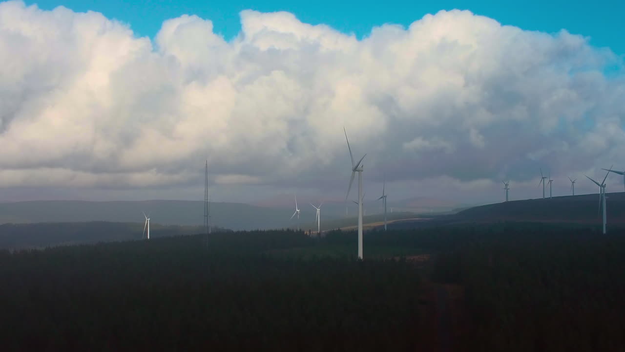 Drone View of Wind Farm in Dense Forest on Mountain Top with Huge Clouds Above. Clean Green Energy Being Generated to Combat Climate Change. Filmed in Wales, UK